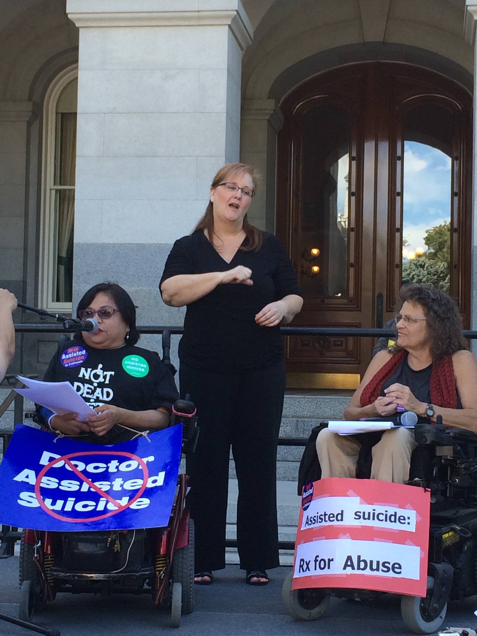 Lillibeth and Marilyn Golden speaking at rally, wearing signs opposing assisted suicide, with an ASL interpreter standing between them.
