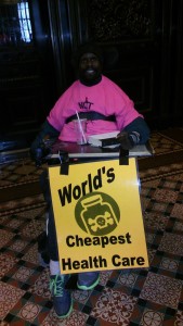 African American wheelchair user in pink NDY tshirt holding yellow poster saying "World's Cheapest Health Care" with a poison bottle depicted.