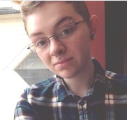 Head and shoulders photo of youthful person with a close cropped haircut, wire rimmed glasses and black, blue and white plaid shirt.