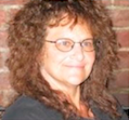 head and shoulders photo of smiling woman with brown curly hair and wire rimmed glasses, a dark top and brick wall behind her