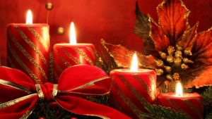 Photo of four lighted red candles with gold glitter, resting on an evergreen bed with a red bow and a poinsettia flower with gold glitter.