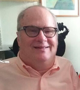 Head and shoulders photo of smiling short haired white man with dark-rimmed glasses wearing a peach colored shirt, with wheelchair headrest and sip-and-puff operating switch visible in the frame.