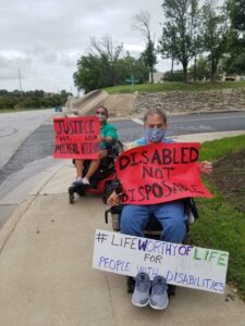 Photo description: Nicky and Ron wearing masks in their wheelchairs holding red signs outside St. David's Hospital in Austin. Nicky's sign reads Justice for Michael Hickson 9-6-1973 to 6-11-2020. Ron's red sign read Disabled Not Disposable and a white sign on his feet Life Worthy Of Life for People With Disabilities