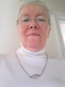 Image of Carol Cleigh Sutton, a fair skinned woman with white hair, pulled back, wire rimmed glasses, and a white turtle neck top with a silver necklace featuring two connected silver handcuffs with small white stones.