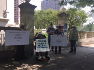 Five protesters with hats and face masks, one in a wheelchair, hold 3 signs in front of stone pillars at the entrance of a walkway toward a building with trees in the background.