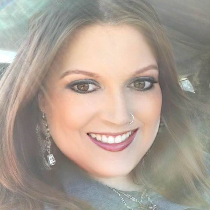Image Description: Headshot of white woman with light brown shoulder length hair, long earrings, a nose ring, and necklace against a light blue collar.