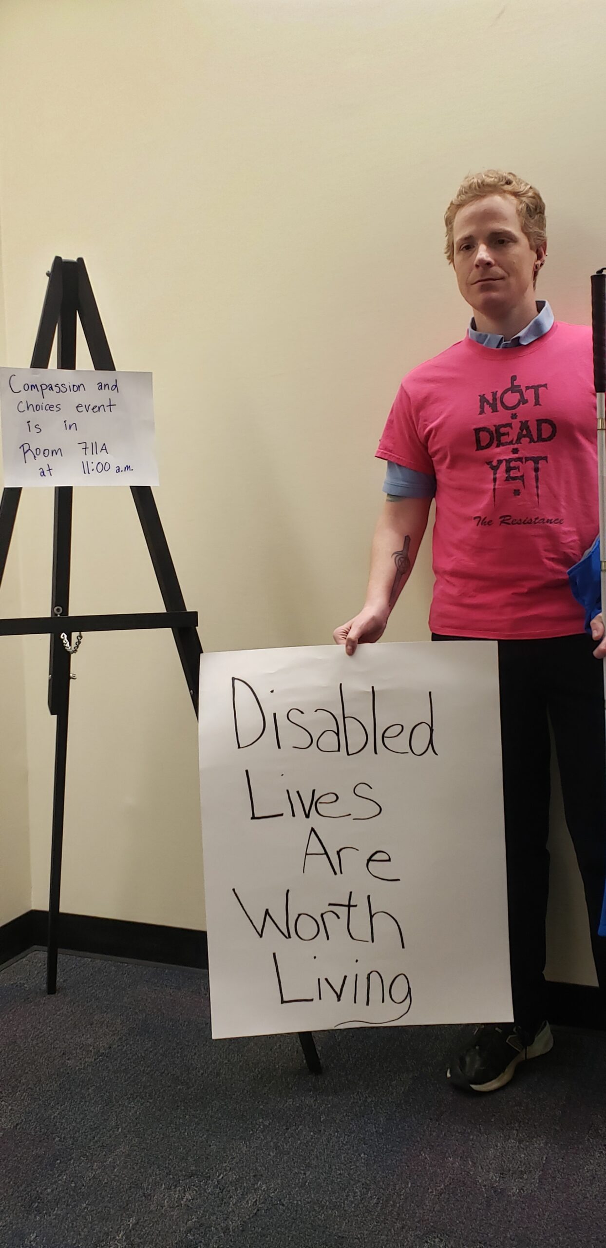 White man with strawberry blond hair wearing a Not Dead Yet t-shirt with black pants, holding a white cane, with a sign saying "Disabled Lives Are Worth Living", near a tripod sign saying "Compassion and Choices event is in Room 711A at 11:00 am".