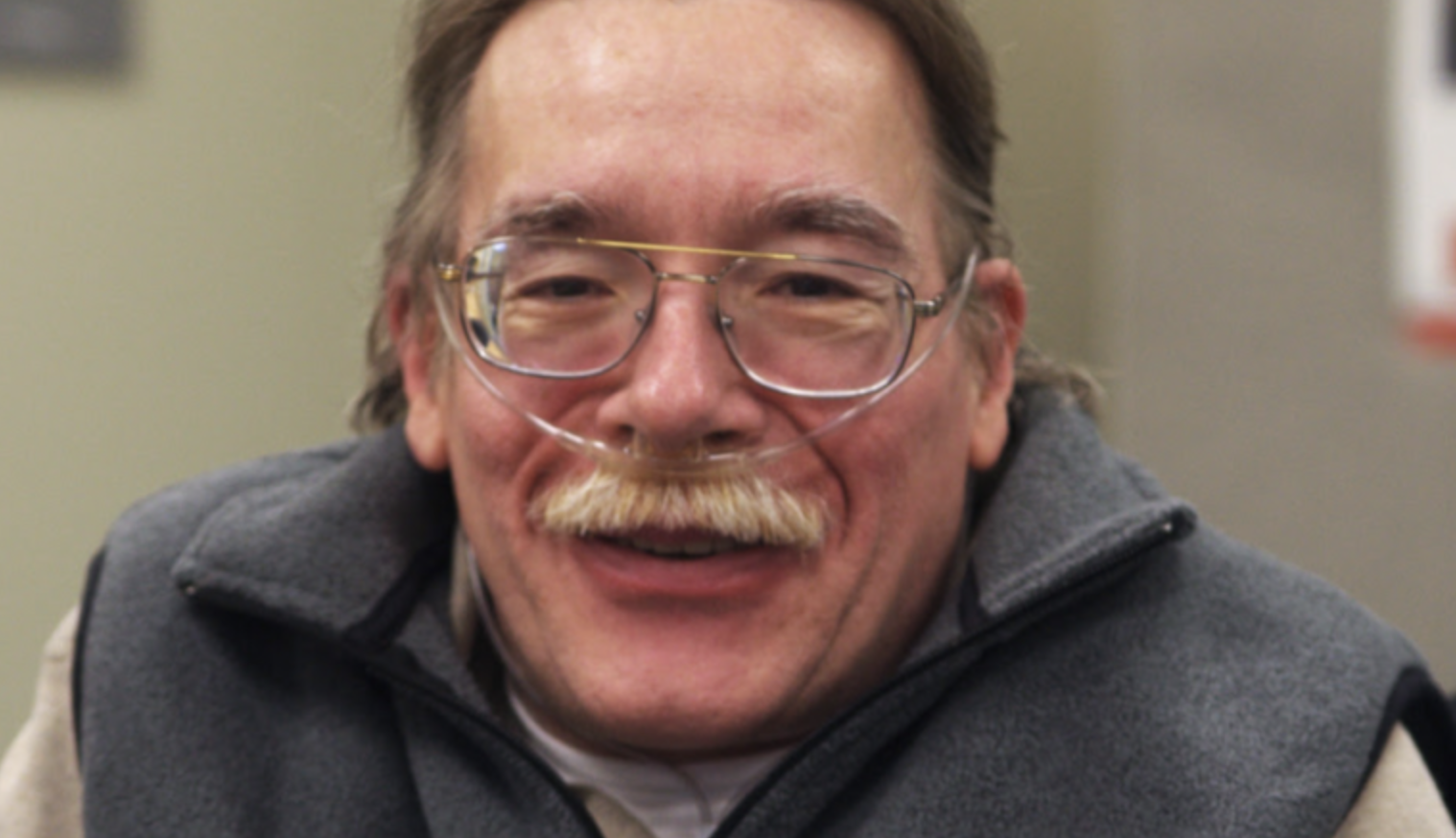 Head and shoulder photo of Paul Spooner, a smiling brown haired white man with a moustache, wire-rimmed glasses, an oxygen nasal canula and a gray fleece jacket.