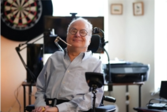 Head and torso photo of white man with short gray hair, glasses and a light blue long sleeved shirt sitting in a motorized wheelchair in a white-walled room with various photos and electronics nearby.