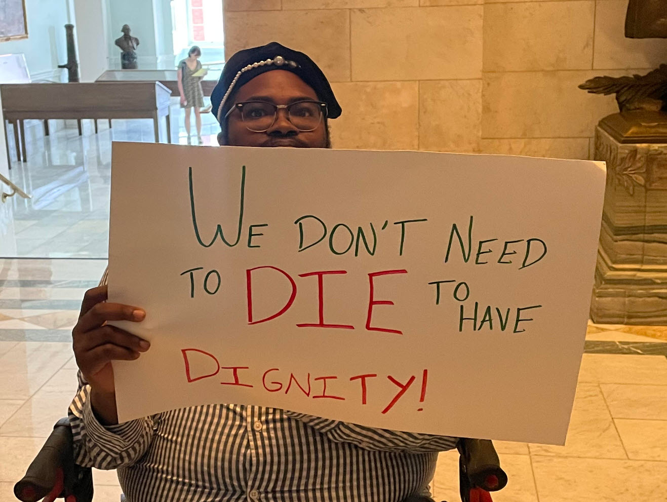 Finn Gardiner, a black man with beard, glasses, and a black hat seated holding up a sign reading "we don't need to die to have dignity."