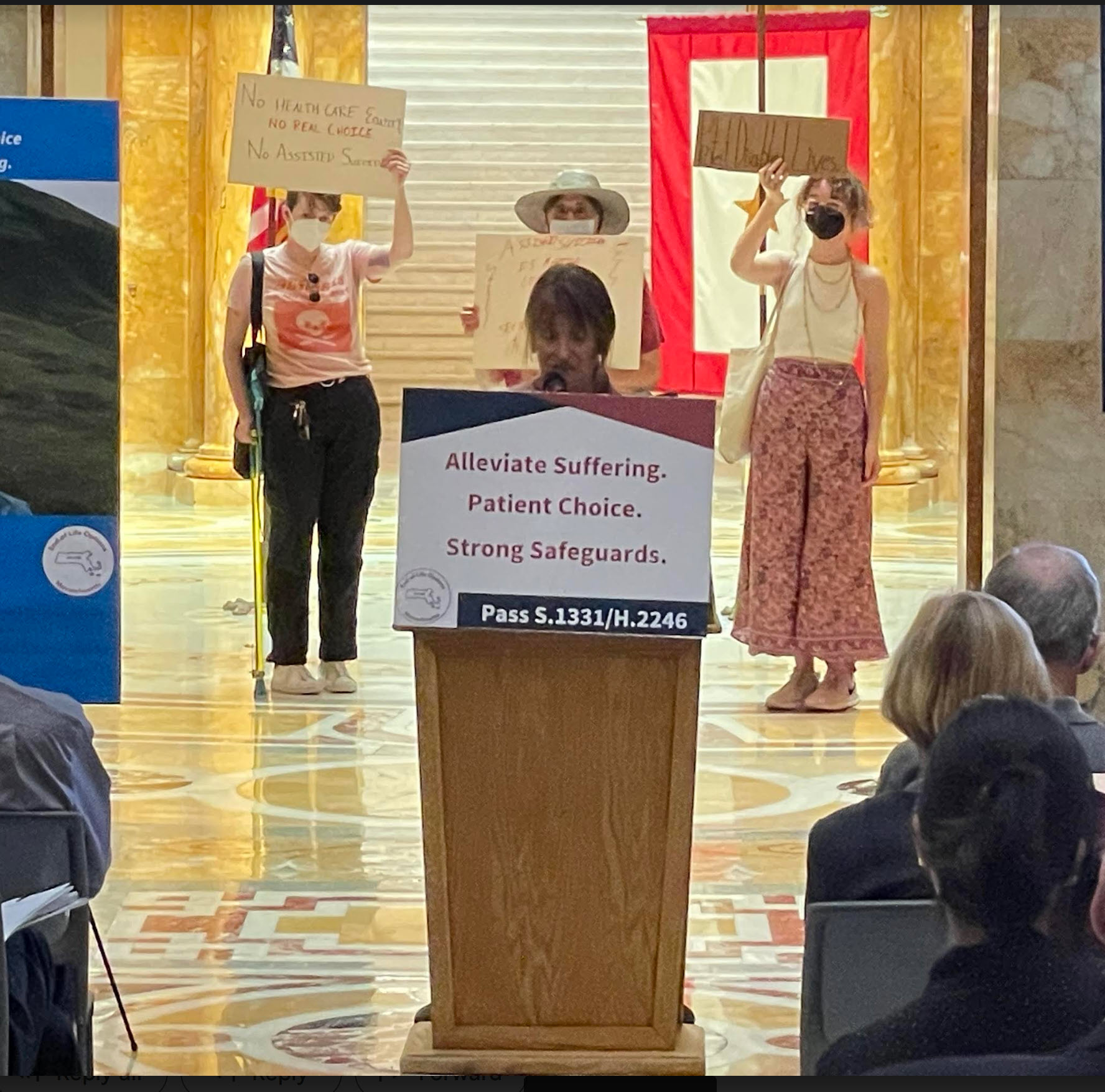 L-R, rear, Jules Good holding sign "No health care equity/No real choice/No assisted suicide.", Don Summerfield, a white man in a broad white hat (partially obscured by speaker at podium in foreground) holding obscured sign, and Jess with sign "protect disabled lives!" Foreground: woman speaking at podium, which has sign reading "alleviate suffering/patient choice / strong safeguards"