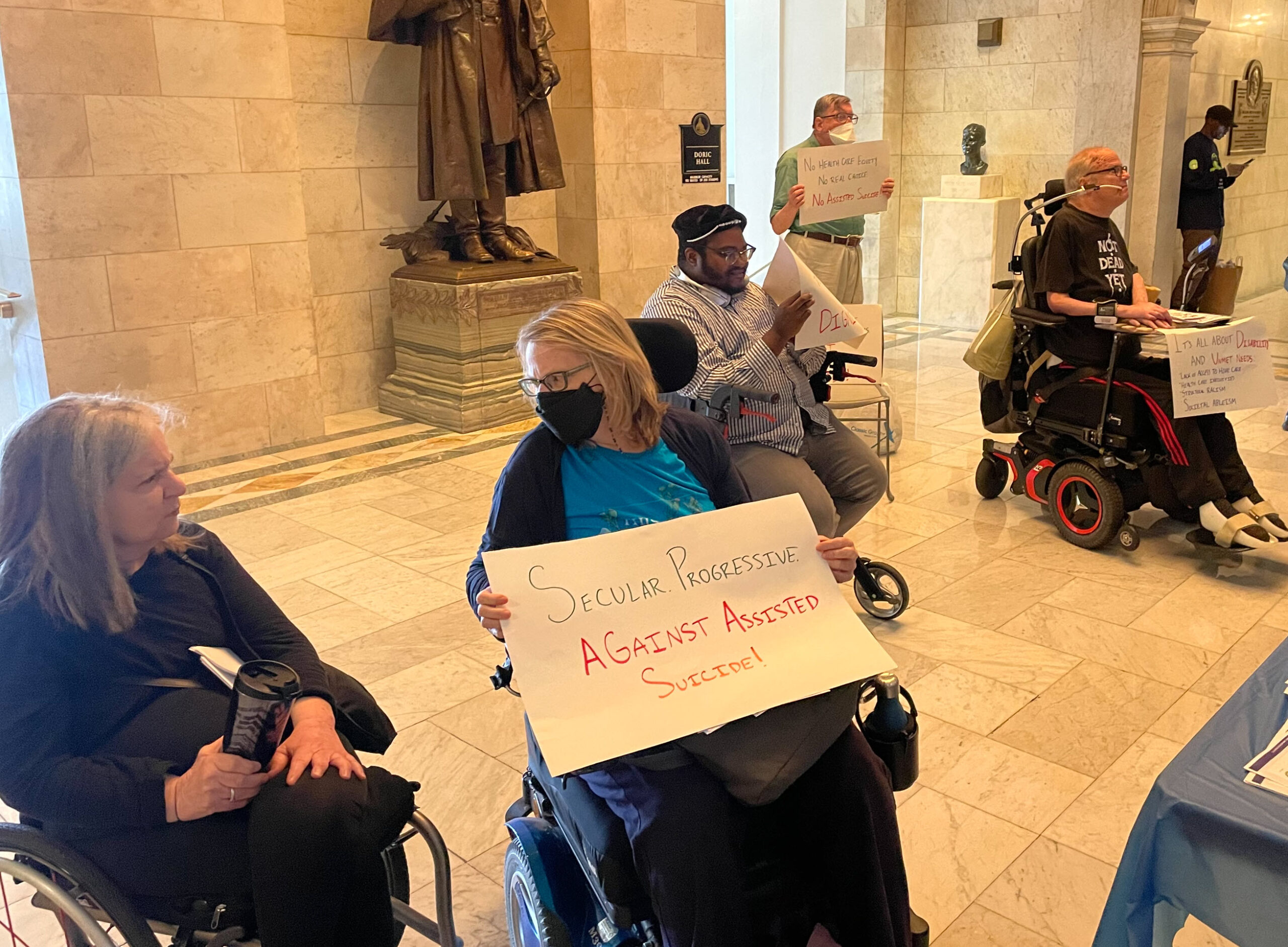Seated Signs: left to right, seated: Pam Daly, a white woman with shoulder length hair in a manual wheelchair Ellen Leigh, a white woman in a power wheelchair with shoulder length hair, a mask and eyeglasses, Finn Gardiner, a black man with beard, glasses and a black hat, John Robinson (standing behind), a white man in a green shirt and light slacks with glasses and a facemask, side-view of John Kelly, a balding white man in a power wheelchair with a "Not Dead Yet" T-shirt and a sign. Ellen's sign reads: "Secular Progressive against Assisted Suicide." John's sign says, "it's all about disability and unmet needs: lack of access to home care / healthcare inequities/structural racism / societal ableism. "