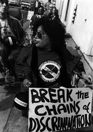 Lillibeth Navarro leading protest march wearing ADAPT accessible transit t-shirt with sign saying "Break the Chains of Discrimination."