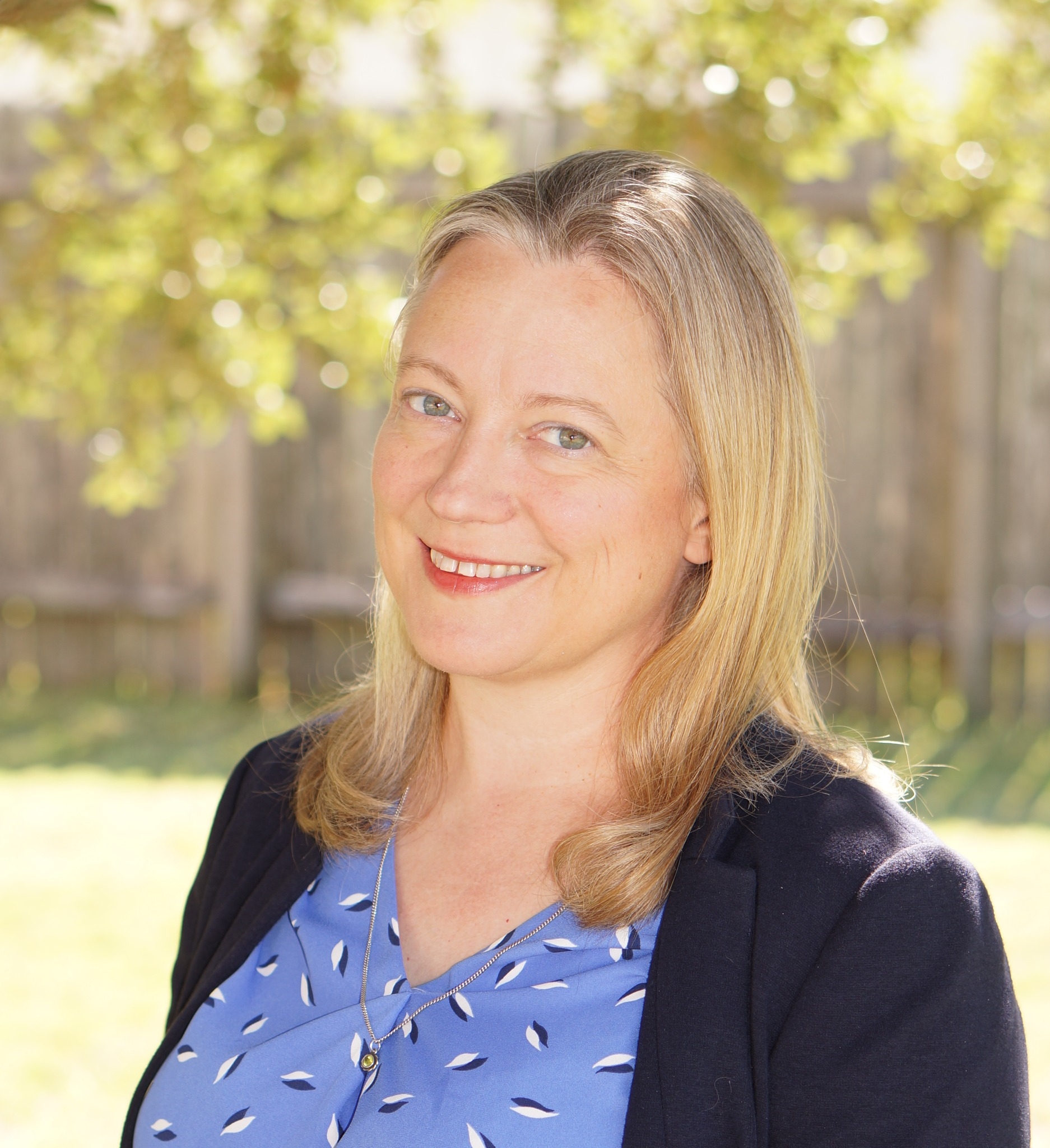 Smiling white woman in her 40s with light-colored hair, wearing a blue suit with trees in the background.