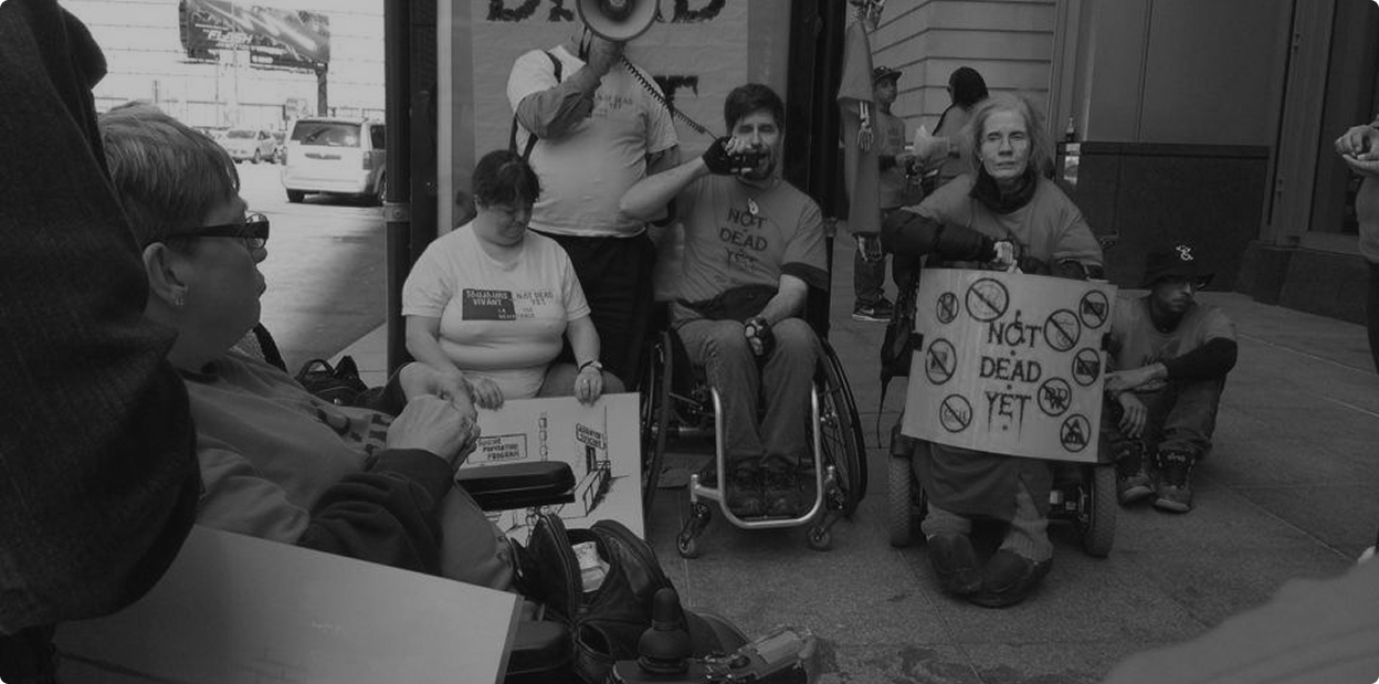 Black-and-white image of Diane Coleman and a group of disabled adults in wheelchairs protesting. One is holding a megaphone. Diane's sign says "Not Dead Yet."