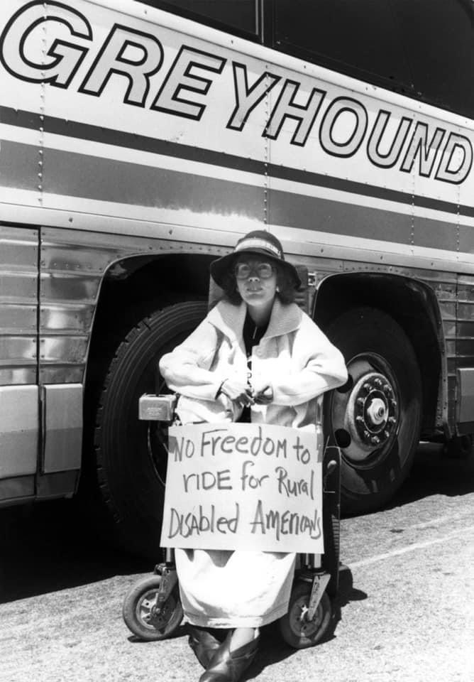 Black-and-white image of Diane Coleman using a mobility scooter in front of a Greyhound bus and holding a sign that reads, “No freedom to ride for rural disabled Americans.”