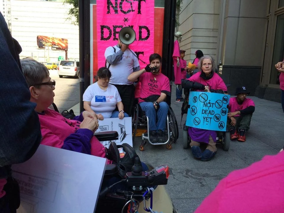 Not Dead Yet activists, including Diane Coleman, several using wheelchairs, gather on a city sidewalk holding pink protest signs. One near the back is holding a megaphone. Diane Coleman is to the right holding a sign that says "Not Dead Yet." Behind the activists is another sign where you can read "Not" and "Dead" in Not Dead Yet.