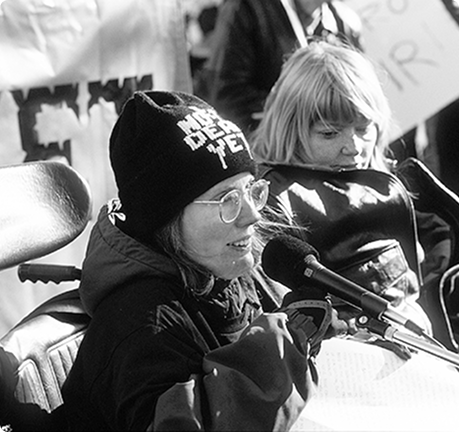 Black-and-white image of Diane Coleman wearing a Not Dead Yet beanie hat and speaking into a microphone. Another activist is next to her.