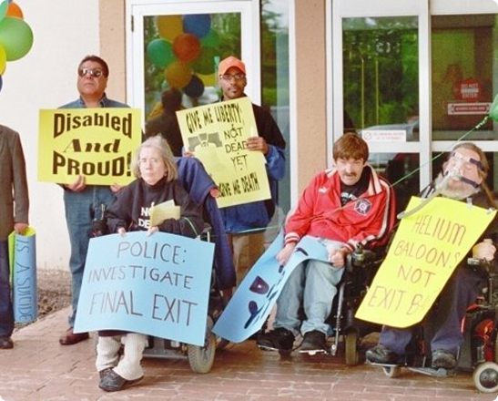 Disability rights activists, including Diane Coleman, several using wheelchairs, hold signs opposing assisted suicide, including “Disabled and Proud” and “Not Dead Yet." Diane Coleman is in black clothing and holding a blue sign labeled "Police: Investigate Final Exit."