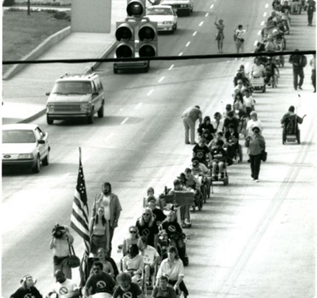 Black-and-white image showing a long procession of disability rights activists, many using wheelchairs, traveling along a city sidewalk. They have an American flag with them. Cars are on the street on the left.