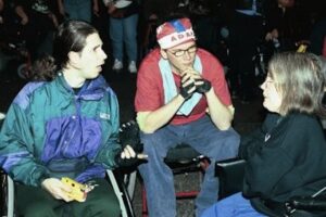 Diane Coleman, sitted in a wheelchair, speaking to two men, both also seated, at an ADAPT gathering while others stand behind them.