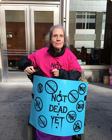 Diane Coleman, a disability rights advocate, in a motor scooter wearing a pink Not Dead Yet shirt and holding a blue protest Not Dead Yet sign.