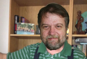Bearded man in a green striped shirt smiles in front of a wooden bookshelf.