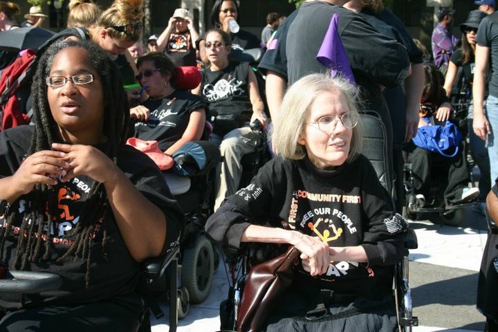 Diane Coleman and another disability rights activist in black shirts that read "Free Our People", sitting in wheelchairs participating in an outdoor demonstration. A large crowd of other disabled activists in black shirts is behind them.