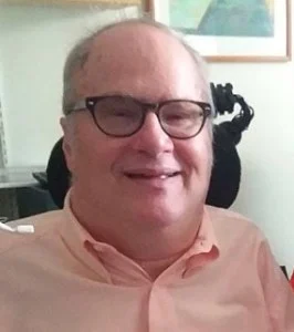 Image of John B. Kelly, a smiling older middle-aged white man with glasses wearing a pink shirt.