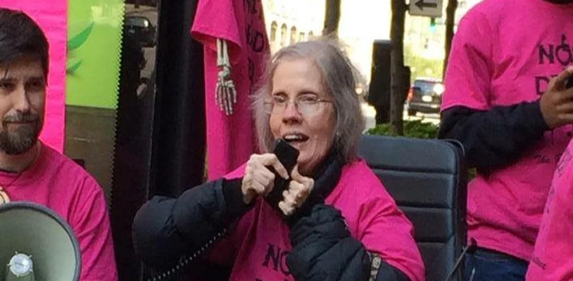 Diane speaking into a microphone in a pink Not Dead Yet shirt. Other disability rights activists are nearby. 