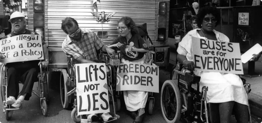 A group of disabled activists, including a young Diane Coleman, protesting. Diane Coleman's sign says "Freedom Rider." The other signs say "Lifts Not Lies," "Buses are for Everyone," and "It's Illegal and a Dog of a Policy." They are all in wheelchairs. 