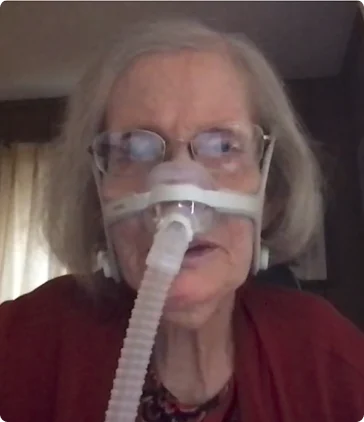 Diane Coleman smiling, wearing a red sweater, glasses, and on oxygen mask over her nose.